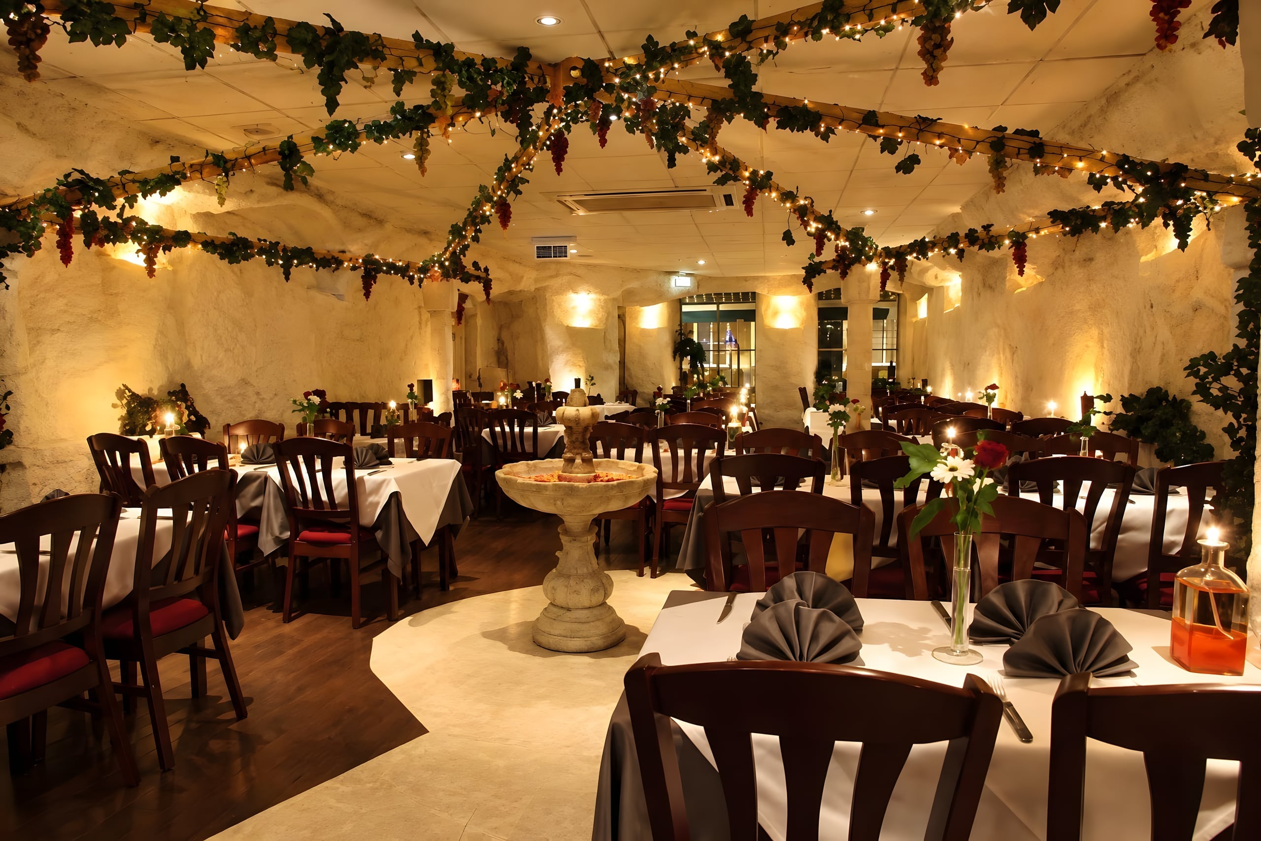 De zaal van Bella Roma bij avond, met kaarslicht, wit gedekte tafels en wijnranken met lichtjes langs het plafond